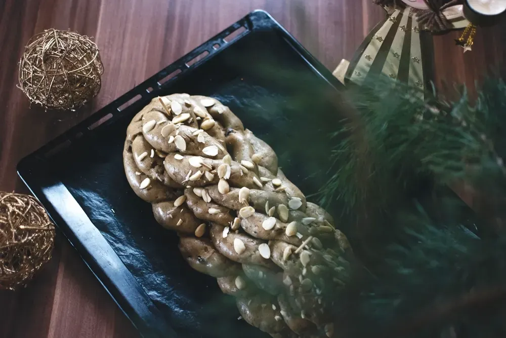 撒着杏仁的辫子形圣诞甜面包放在黑色烤盘上，旁边是松枝和装饰